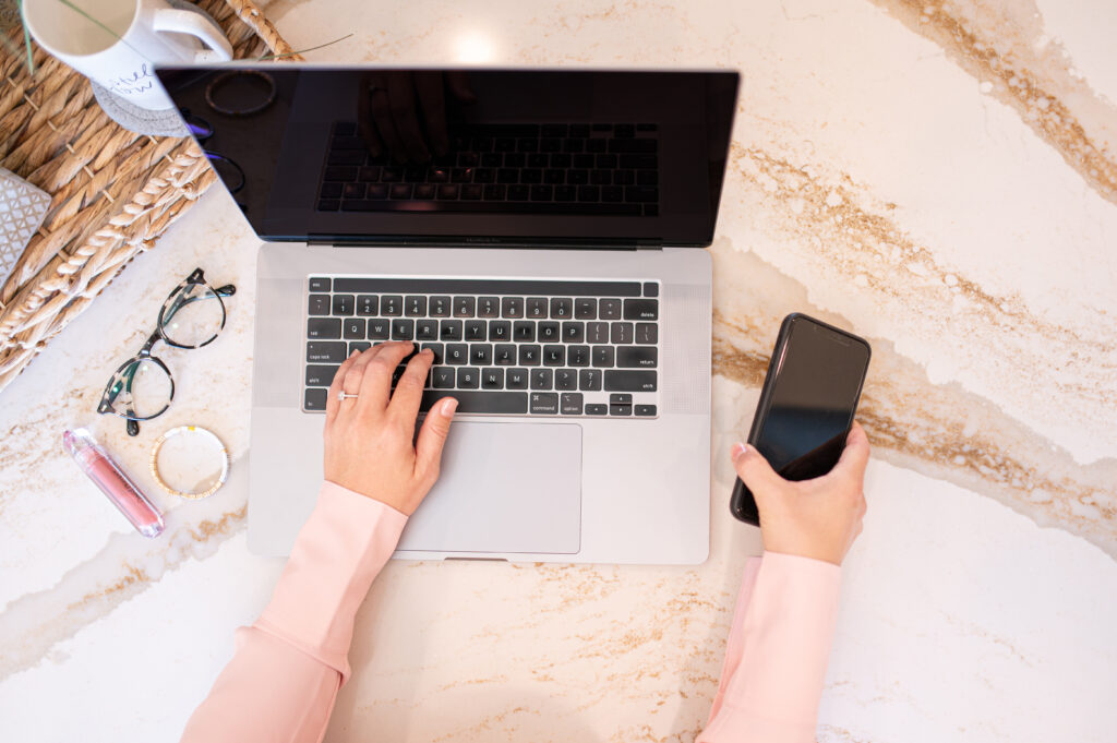 Flat lay of a wedding professional working on a laptop with a smartphone in hand, styled desk with glasses, lip gloss, bracelet, and coffee mug — representing blogging and SEO for wedding businesses.